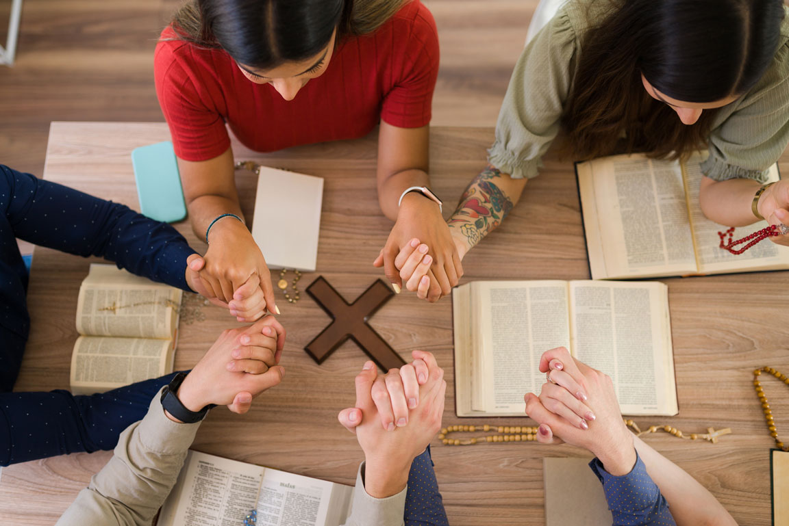 high-angle-catholic-young-men-women-holding-each-other-hands-while-praying-together-around-table-with-christian-cross
